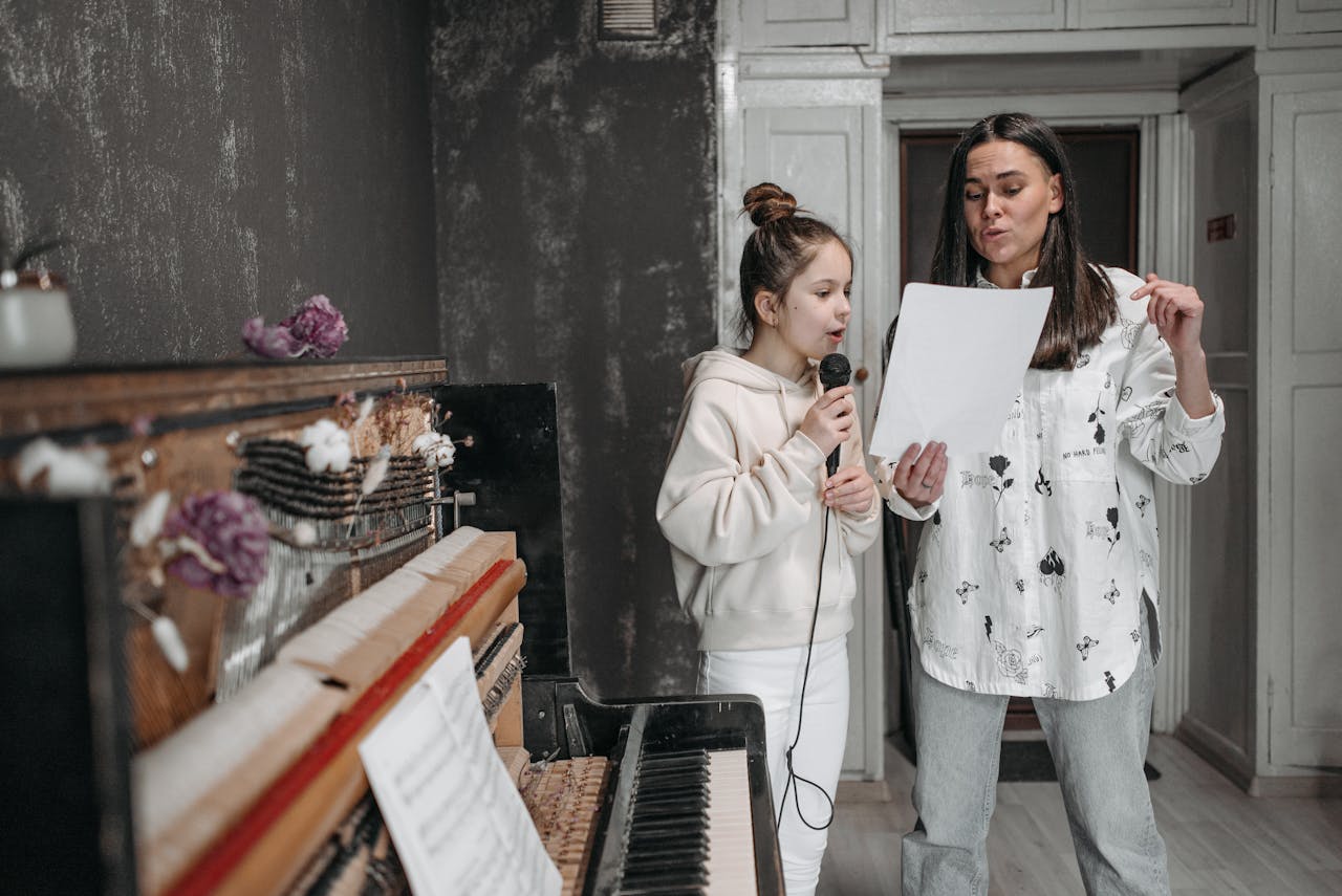 A girl taking a voice lesson from her teacher, singing into a microphone by a piano.